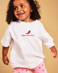 A toddler girl with curly dark hair is wearing a white oversized t-shirt with a red chili pepper graphic and the text 'SMALL BUT SPICY' on the chest, paired with pink shorts featuring a red star pattern. She is posing against a solid warm beige background.