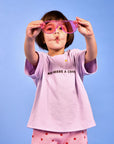 A young child wearing a light purple t-shirt with 'MUM NEEDS A COFFEE' text and pink shorts with red stars, holding pink novelty sunglasses in front of their face against a solid blue background.
