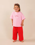 A young child standing barefoot against a plain beige studio background, wearing an oversized light pink T-shirt with SMALL BUT SASSY red text and a red heart on the chest, paired with bright red pants.