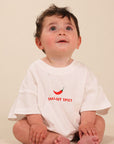 A baby sitting cross-legged wearing a white short-sleeve t-shirt with a small red chili pepper graphic and the text 'Small but SPICY' printed on the chest, set against a plain beige background.