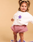 A toddler girl with pigtails stands on a purple star-shaped mat against a beige studio background. She is wearing a white graphic T-shirt with a purple star and the words 'I AM A STAR.
