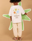 A child stands with their back to the camera wearing a white T-shirt with a colorful star graphic and 'TAURUS energy' text, paired with cream pants, against a solid beige background.