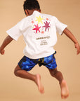 A child jumps in the air wearing a white Aries t-shirt with colorful star graphics on the back and blue patterned trunks, set against a warm beige background with a red star-shaped chalk outline on the floor.