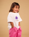 A young girl with shoulder-length brown hair smiling and laughing, wearing a white short-sleeve t-shirt with a purple star graphic and the text 'I'M A STAR' in pink pants, posed against a solid beige studio background.