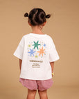 A young girl stands with her back to the camera wearing a white graphic T-shirt and pink gingham shorts, standing on a red star-shaped rug against a solid beige background.