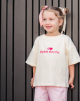 Child wearing a white t-shirt with berry boujee and pink pants standing against a dark vertical panel background