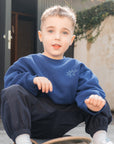 Child sitting on a skateboard wearing a blue sweatshirt and dark pants.