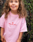 A child with curly hair is sticking out their tongue while wearing a pink t-shirt with 'SMALL BUT SASSY' embroidered in red on the front.