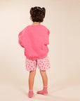A young child viewed from behind, wearing a bright pink oversized sweatshirt, pink shorts with small red heart patterns, and pink-and-white striped socks, standing against a plain beige studio background.