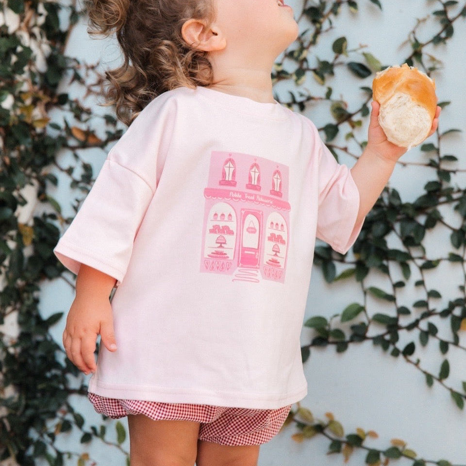 Child wearing a pink t-shirt with a graphic design, standing against a wall with greenery.