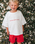 Child wearing a white t-shirt with eat sleep snack repeat and red shorts standing against a leafy background