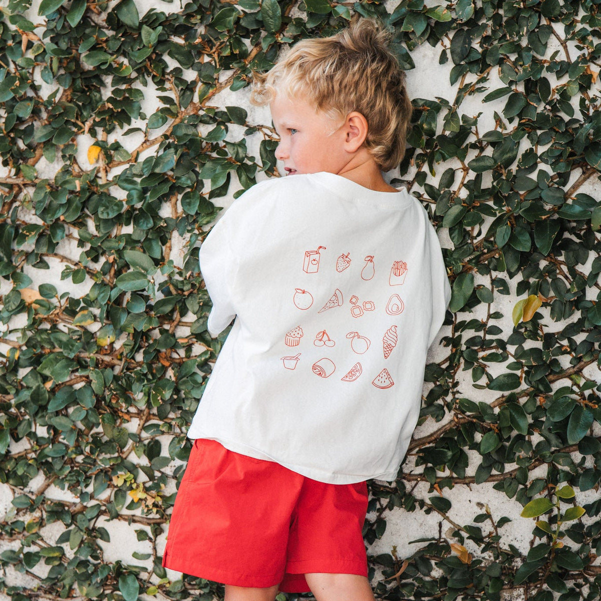 Child wearing a eat sleep snack repeat tee with favourite food patterns and red shorts standing against a leafy wall.