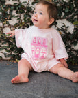 Baby wearing a pink patisserie bakery design, sitting on a stone surface with a leafy background.