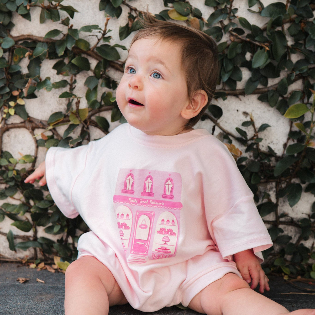Baby sitting with a pink patisserie bodysuit against a leafy background.
