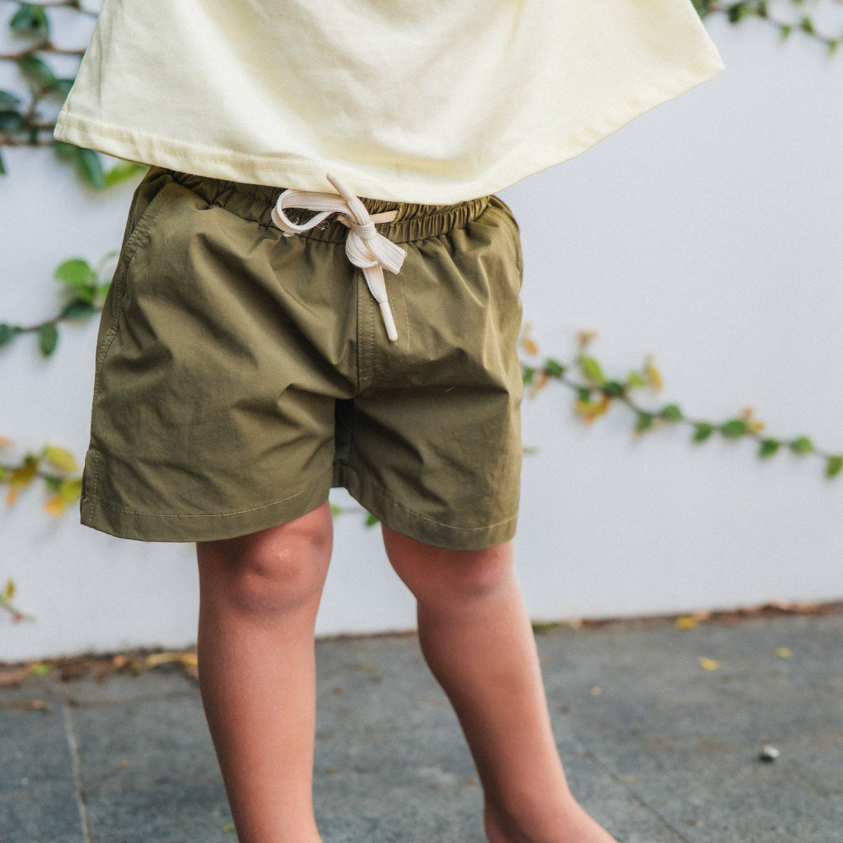 Child wearing green shorts and a yellow top against a white wall with greenery.