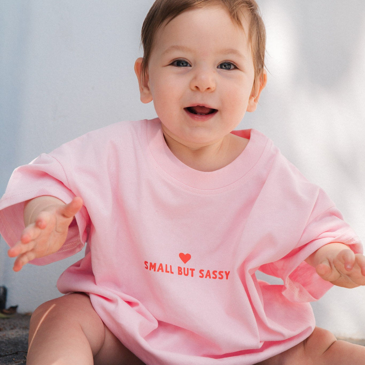 Baby wearing a pink bodysuit with 'Small But Sassy' text sitting on a floor.