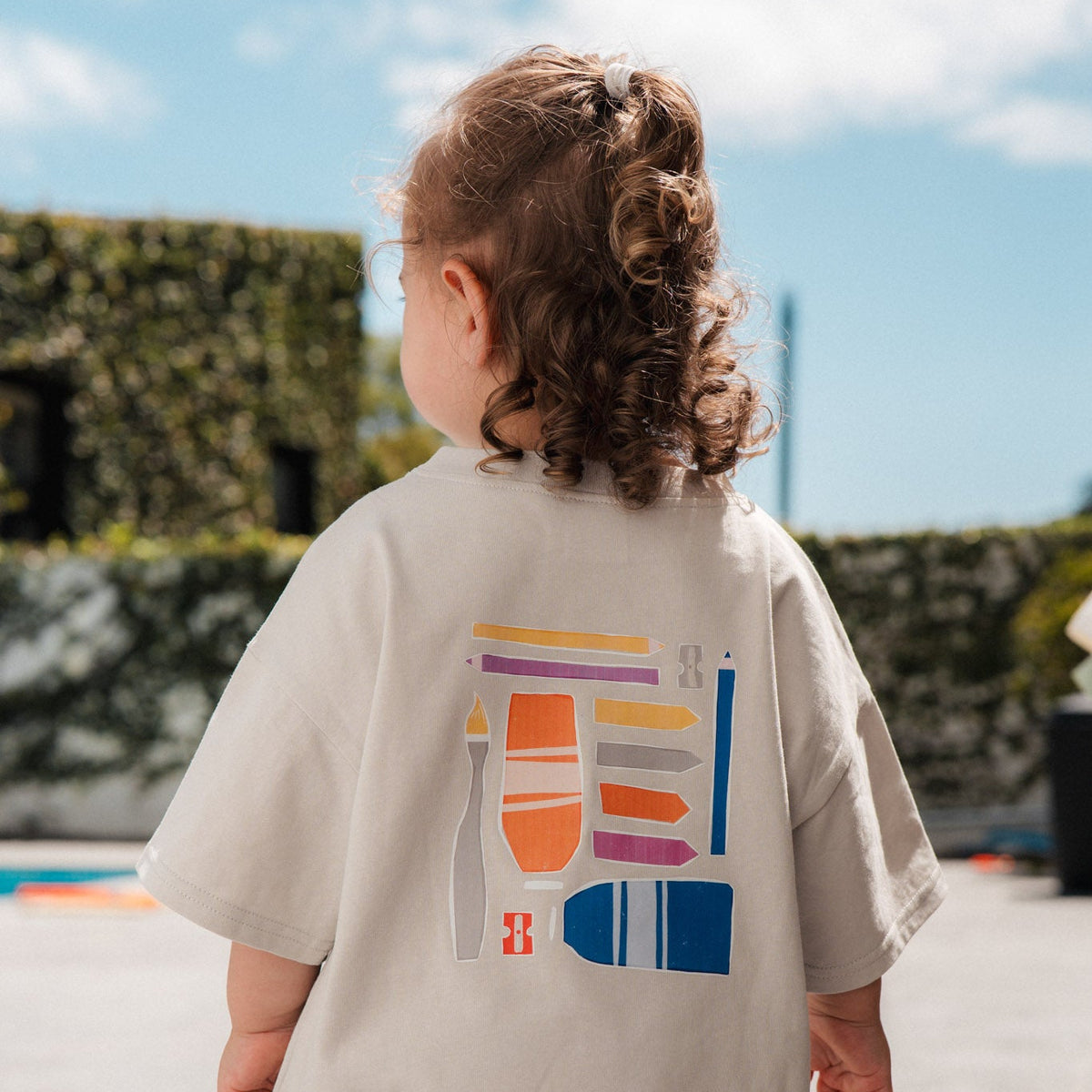 Child wearing a beige dress with a colorful graphic on the back, standing outdoors.