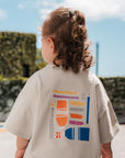 Child wearing a beige dress with a colorful graphic on the back, standing outdoors.