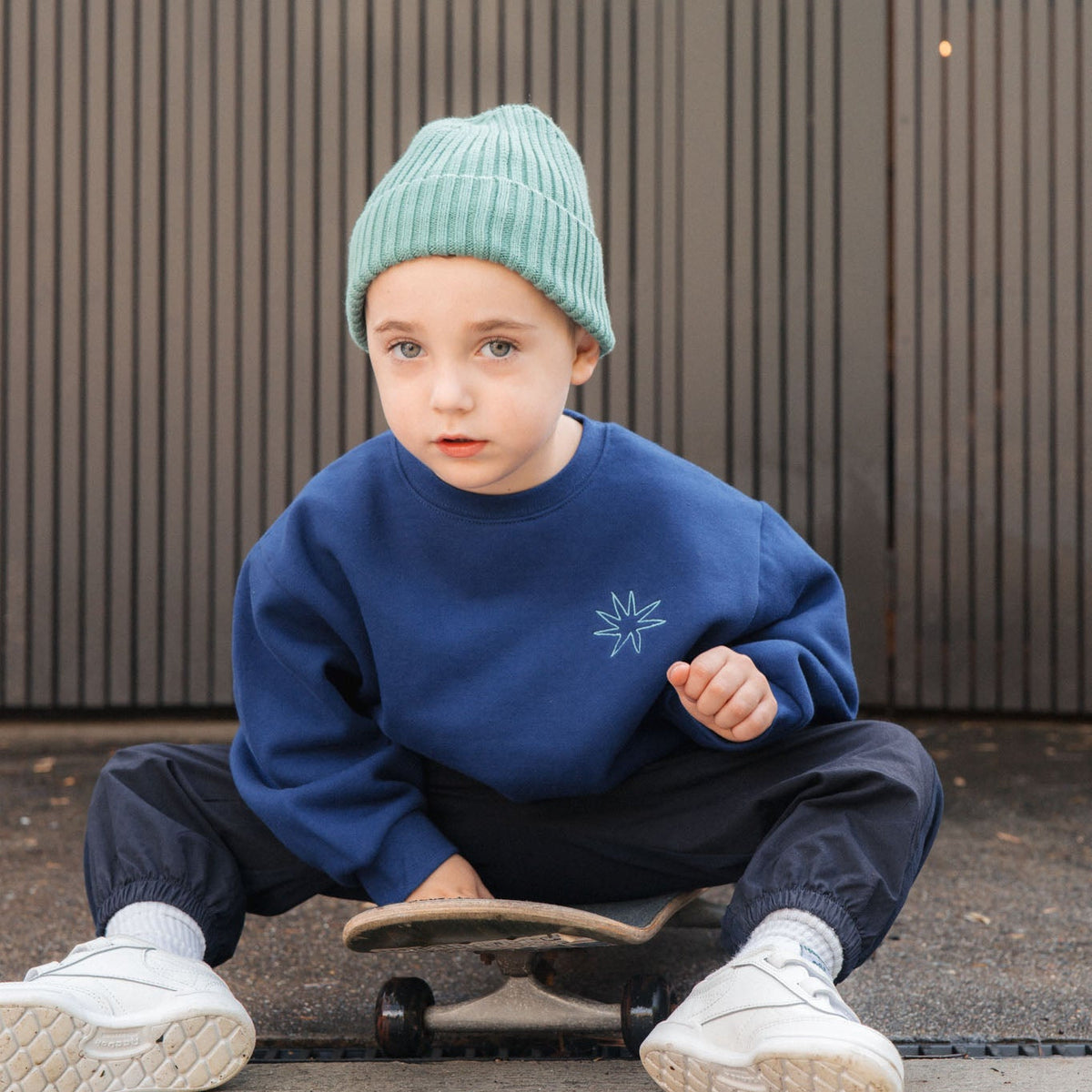 Child wearing a blue sweatshirt and green beanie sitting on a skateboard.