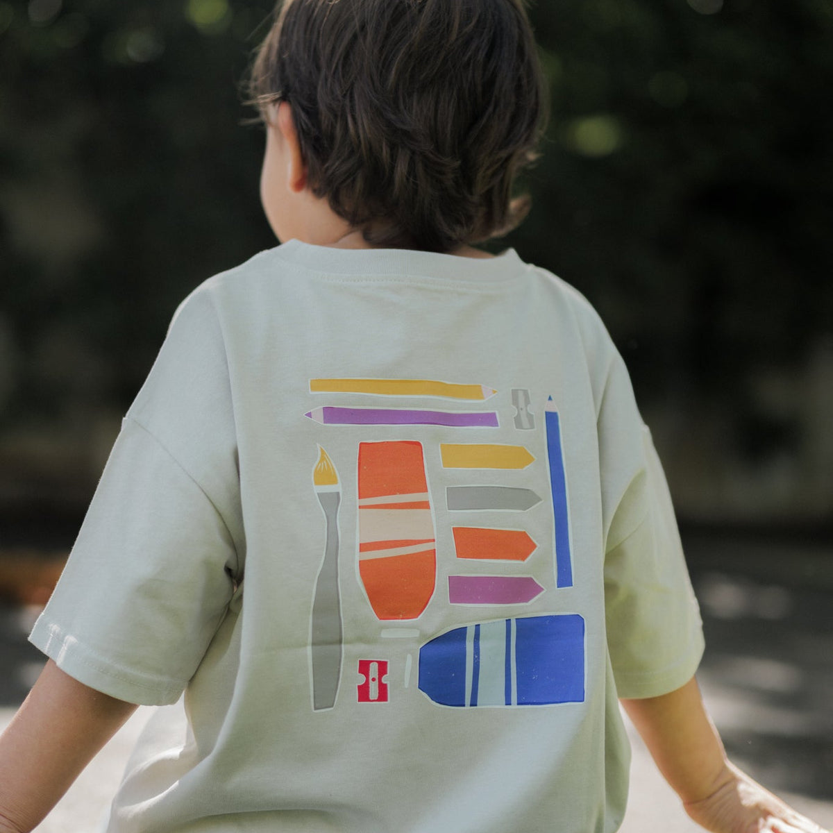 Child wearing a art t-shirt with colorful geometric design sitting on grass.