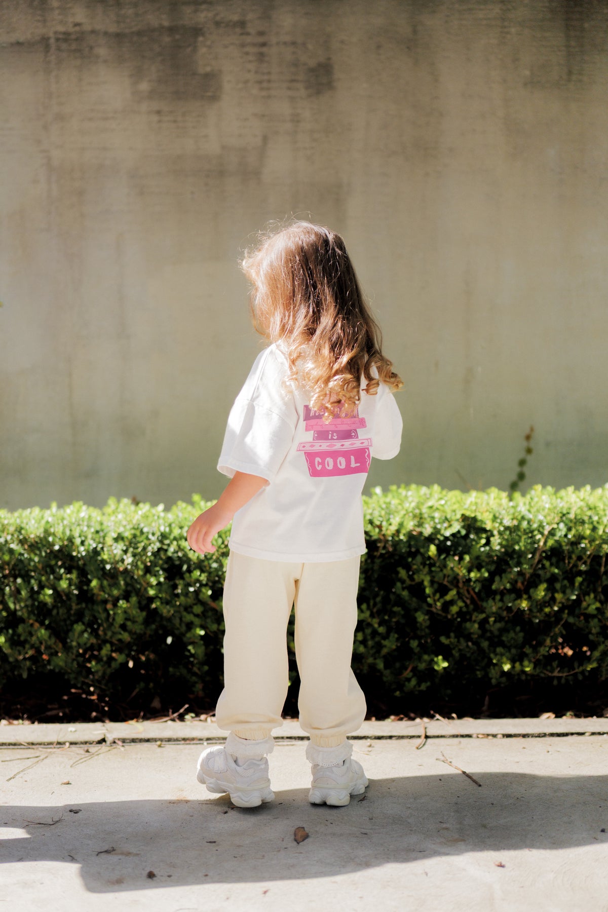 Child wearing a white shirt with a pink design and beige pants standing in front of a concrete wall and green bushes.