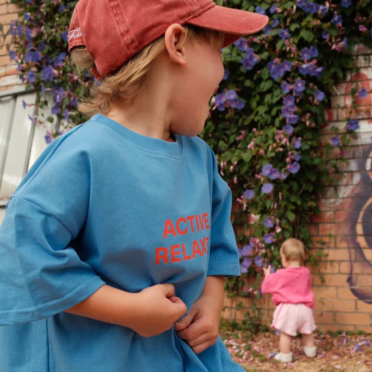 Child wearing a blue shirt with 'ACTIVE RELAXER' text, standing outdoors with a brick wall and flowers in the background.