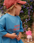Child wearing a blue shirt with 'ACTIVE RELAXER' text, standing outdoors with a brick wall and flowers in the background.