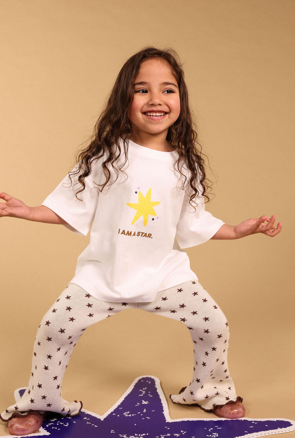 A young girl with long curly brown hair poses in a white t-shirt and matching star-patterned pajama pants, standing on a purple star-shaped rug against a solid beige background.