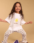 A young girl with long curly brown hair poses in a white t-shirt and matching star-patterned pajama pants, standing on a purple star-shaped rug against a solid beige background.