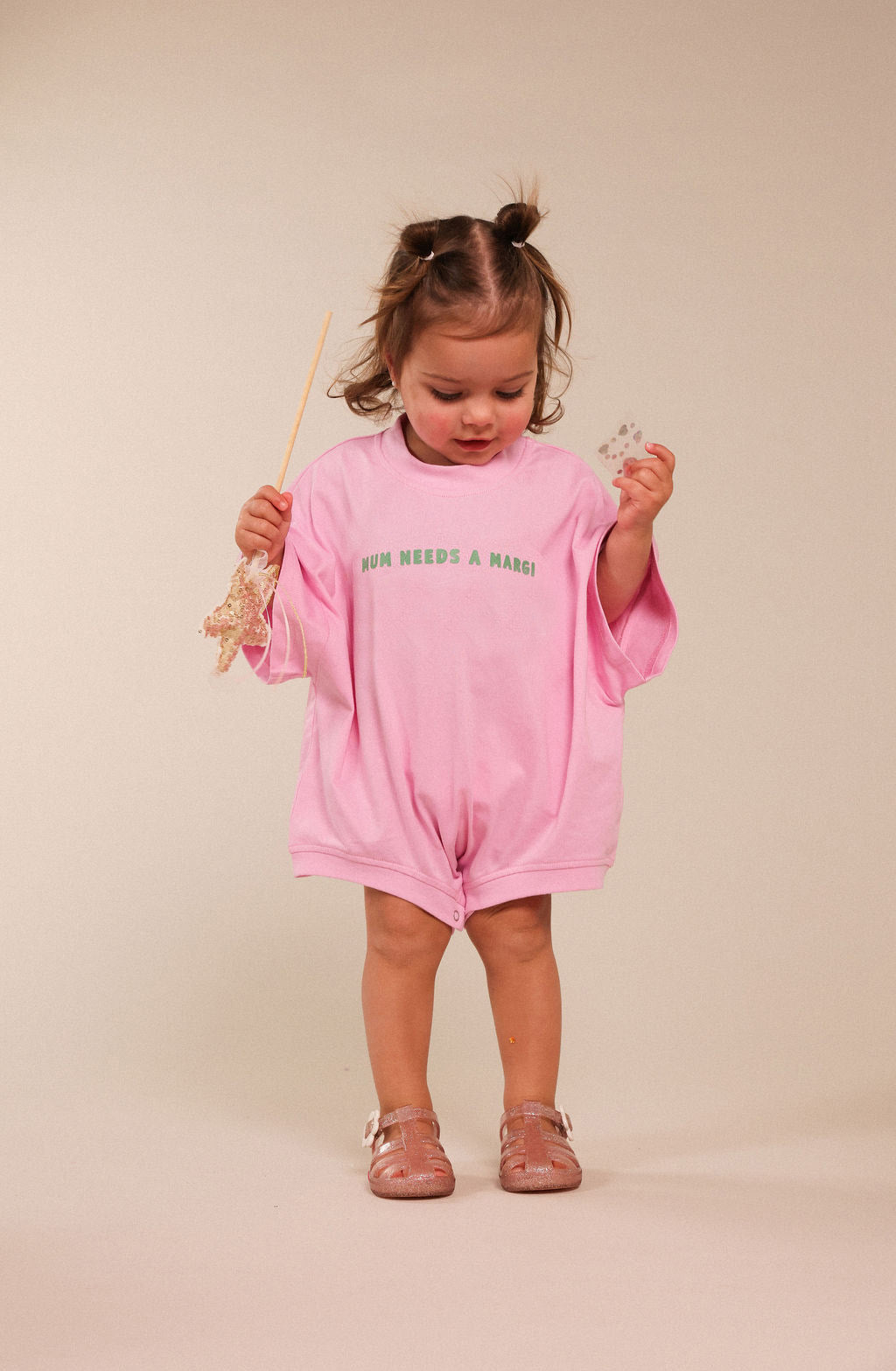 A toddler girl stands against a plain beige studio background wearing a pink short-sleeve romper with green text reading 'MUM NEEDS A MARGI!' across the chest. She holds a wand in one hand and a small object in the other, wearing pink sandals.
