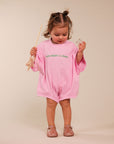 A toddler girl stands against a plain beige studio background wearing a pink short-sleeve romper with green text reading 'MUM NEEDS A MARGI!' across the chest. She holds a wand in one hand and a small object in the other, wearing pink sandals.