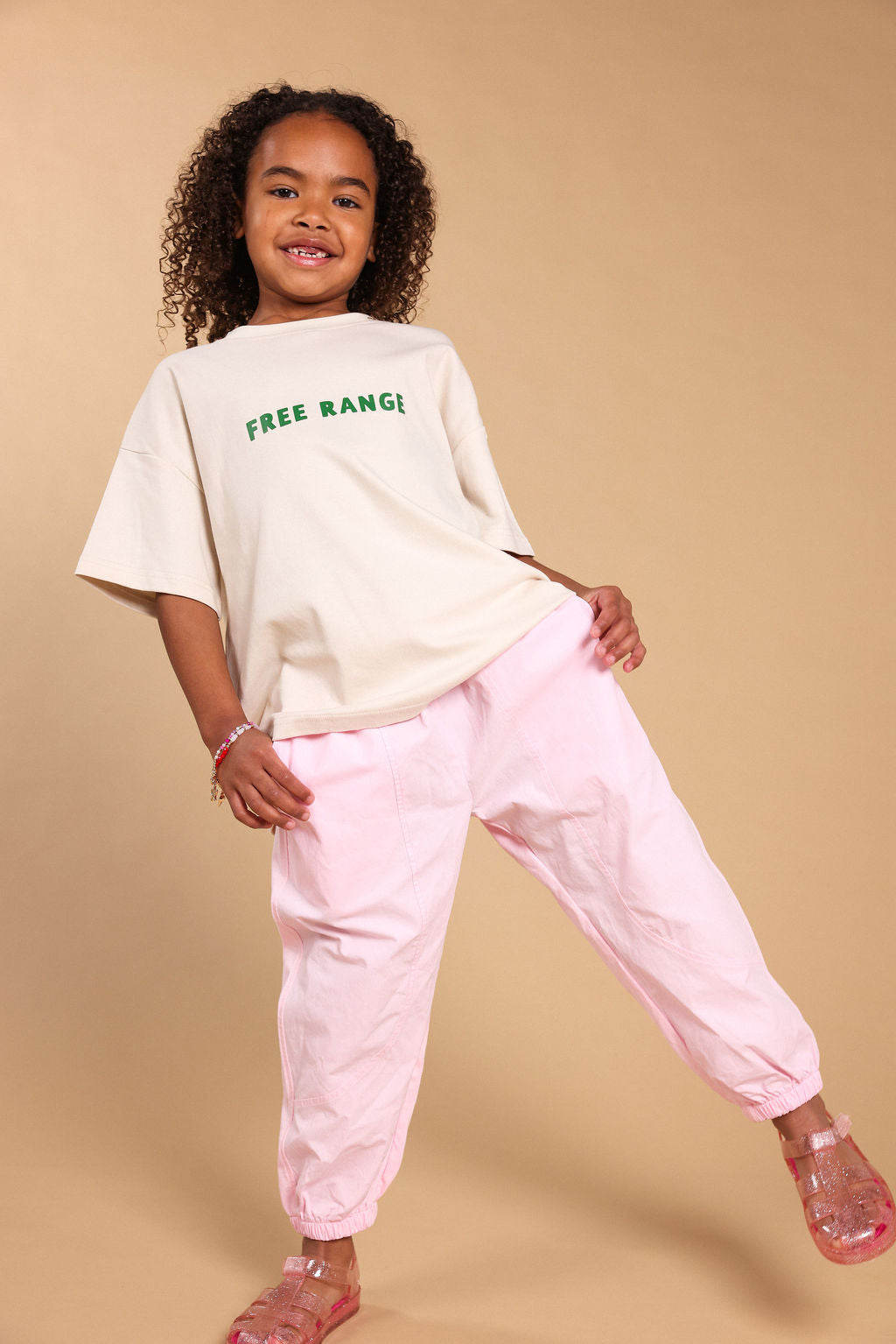 A young girl with curly hair is modeling a cream-colored oversized t-shirt with green 'FREE RANGI' text and light pink baggy pants, standing against a solid beige studio background.