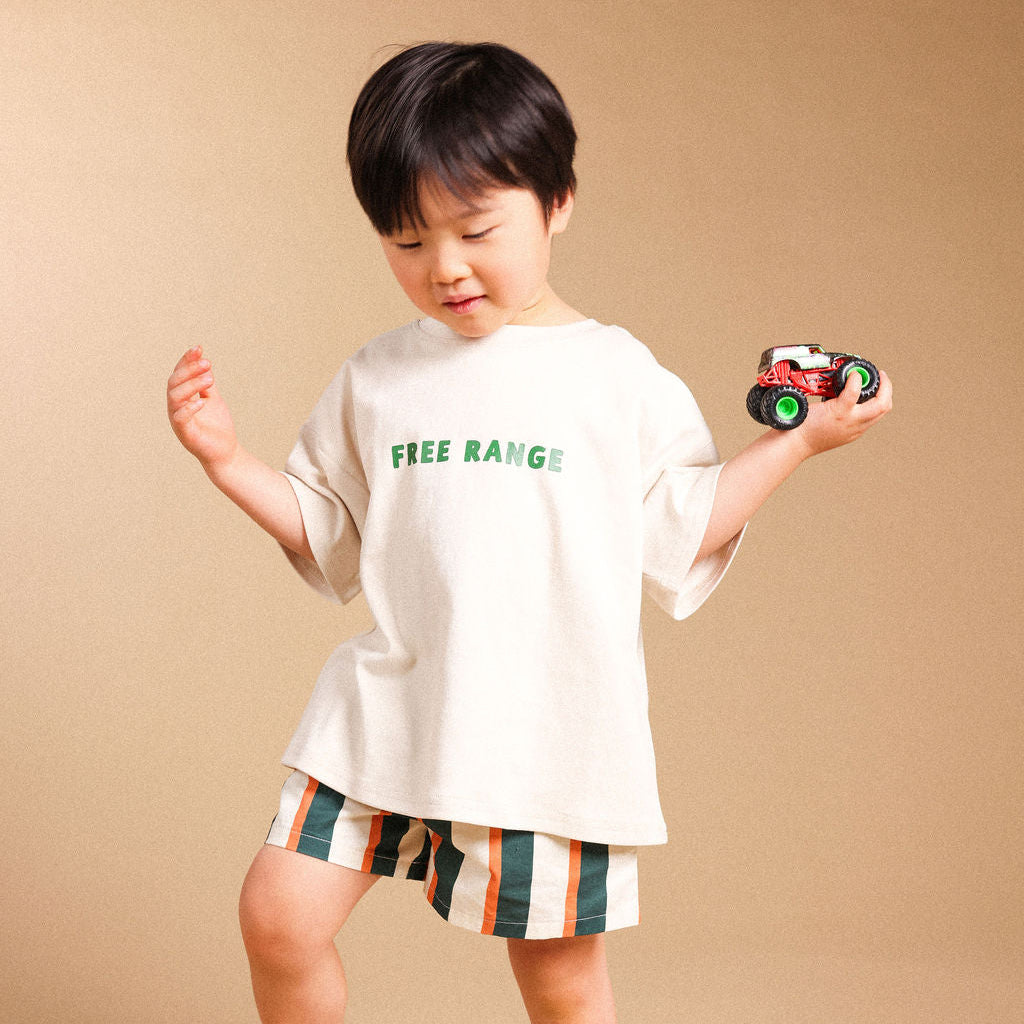 A toddler boy wearing a beige oversized t-shirt with green 'FREE RANGE' text and striped shorts, holding a small toy truck against a solid beige studio background.
