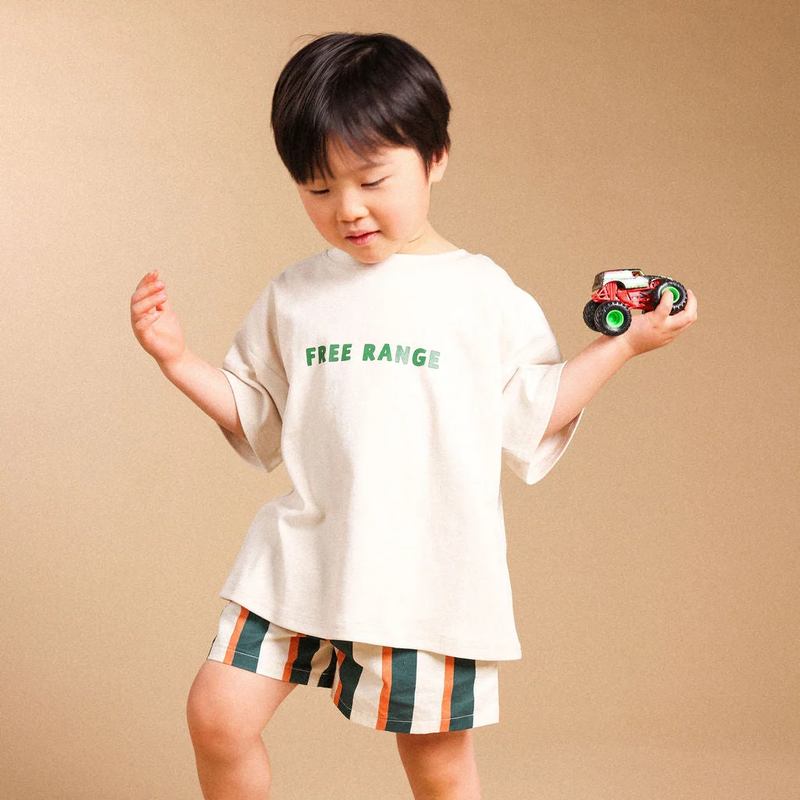 A toddler boy wearing a beige oversized t-shirt with green 'FREE RANGE' text and striped shorts, holding a small toy truck against a solid beige studio background.