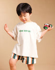 A toddler boy wearing a beige oversized t-shirt with green 'FREE RANGE' text and striped shorts, holding a small toy truck against a solid beige studio background.