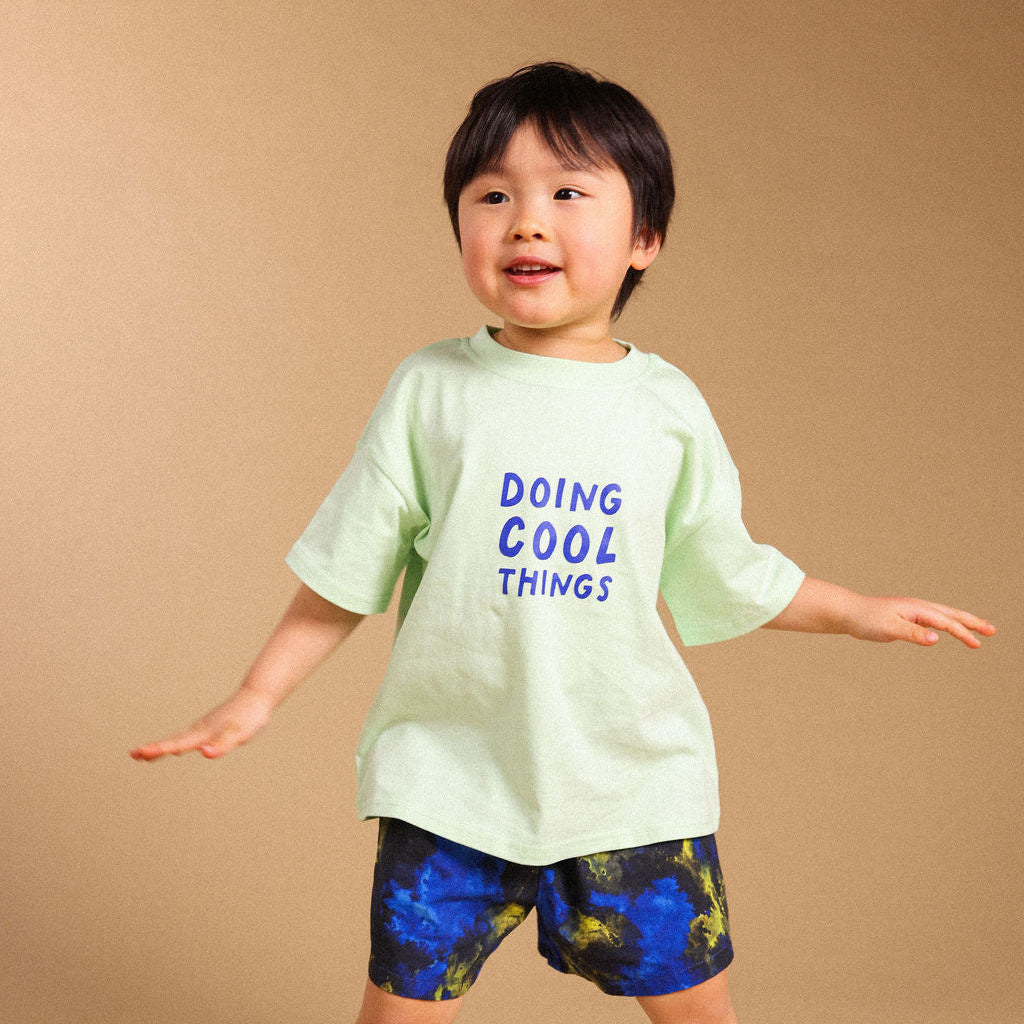 A young child modeling a light green t-shirt with blue text that reads 'DOING COOL THINGS' and blue patterned shorts, standing against a solid beige background.
