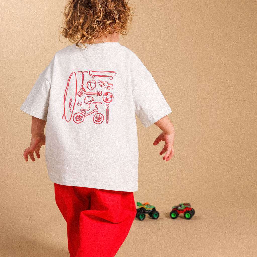 A child seen from behind wearing a white oversized T-shirt with a red graphic print on the back and bright red pants, standing against a plain beige studio background.
