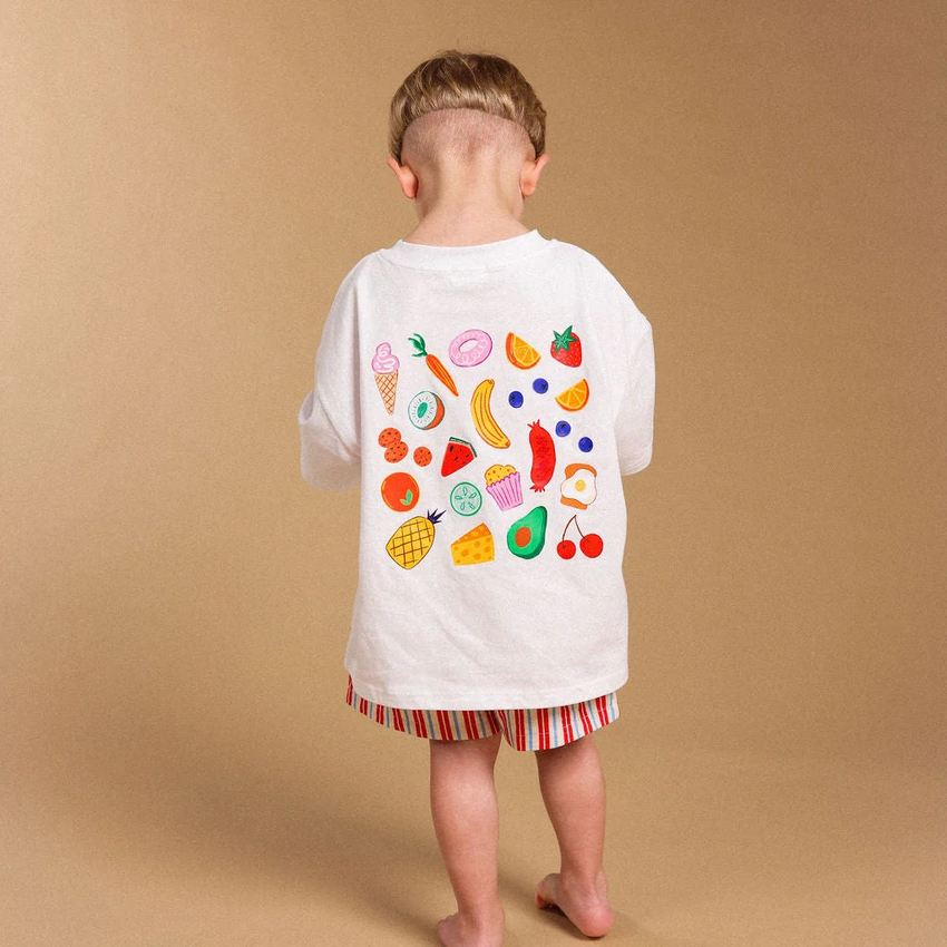 A child standing with their back to the camera, wearing a white t-shirt with a colorful fruit and vegetable pattern on the back, and red, orange, and white striped shorts, against a solid tan studio backdrop.