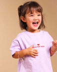 A young girl with dark hair in a ponytail is smiling and laughing while wearing a light purple t-shirt with red text that reads 'SMALL BUT SASSY' and a small red heart, paired with cream shorts decorated with small brown stars. She is standing against a solid beige studio background.