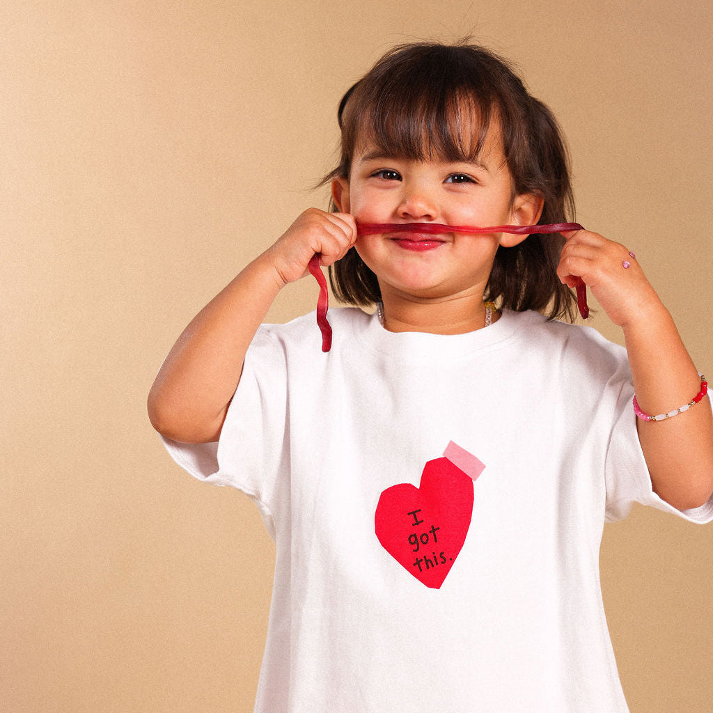 A young girl with dark hair wearing a white t-shirt with a red heart graphic that reads 'I got 2013' and pink pants, holding a red string horizontally in front of her mouth against a solid beige background.
