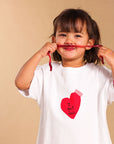 A young girl with dark hair wearing a white t-shirt with a red heart graphic that reads 'I got 2013' and pink pants, holding a red string horizontally in front of her mouth against a solid beige background.