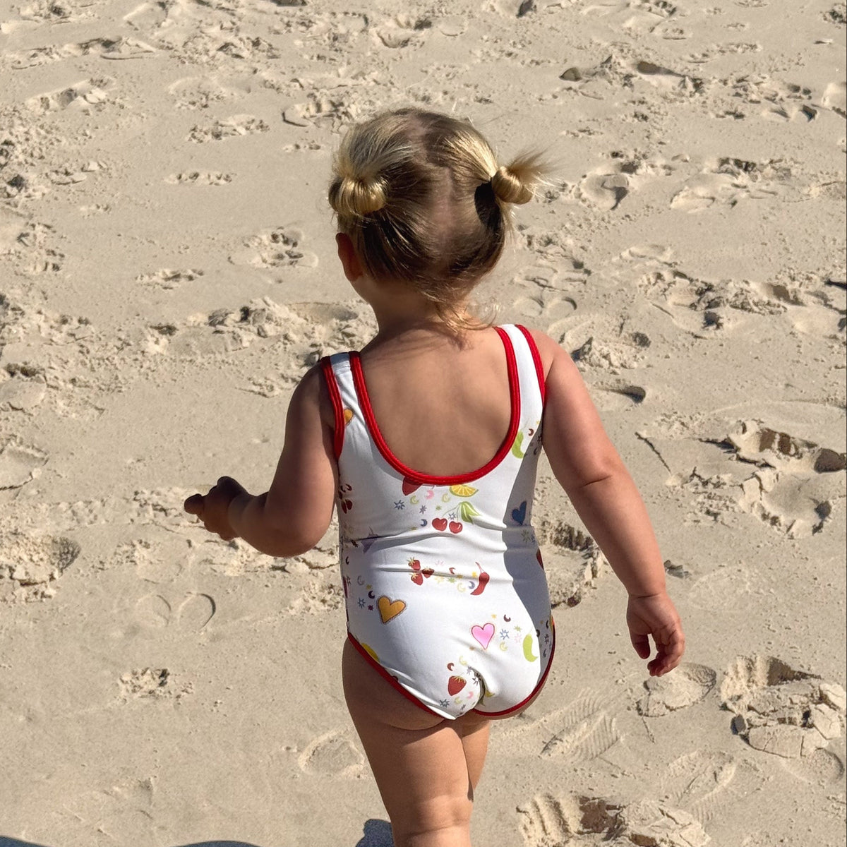 A toddler walking on a sandy beach from behind, wearing a white one-piece swimsuit with red trim and a small multicolor fish pattern.