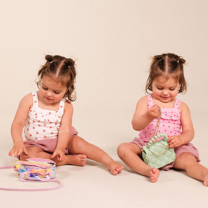 Two young toddlers sitting on a light beige studio floor, playing with small toy bags. The child on the left wears a white sleeveless top with pink hearts and pink shorts, interacting with a clear bag containing colorful items and a pink strap.