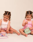 Two young toddlers sitting on a light beige studio floor, playing with small toy bags. The child on the left wears a white sleeveless top with pink hearts and pink shorts, interacting with a clear bag containing colorful items and a pink strap.