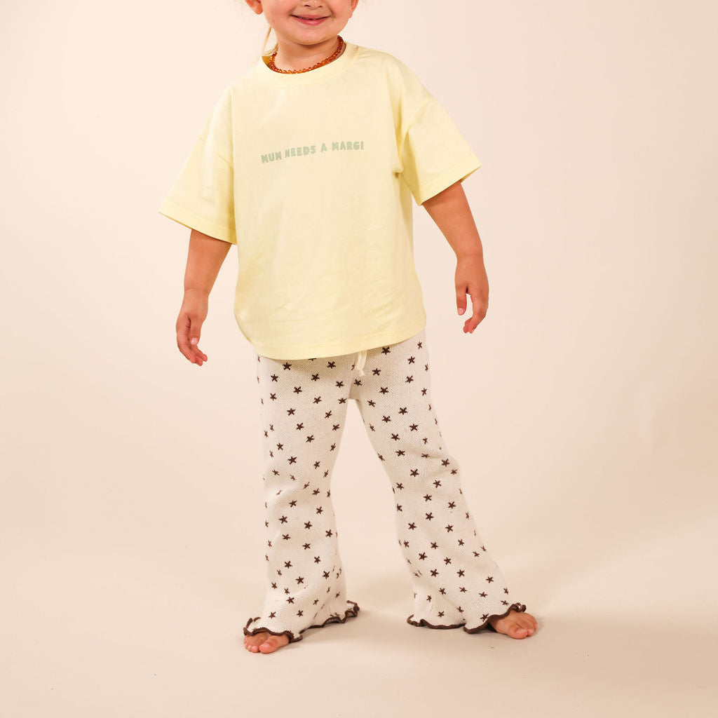 A young child modeling a pale yellow short-sleeve t-shirt with green text and white pants with black polka dots, standing against a solid beige studio background.