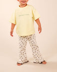 A young child modeling a pale yellow short-sleeve t-shirt with green text and white pants with black polka dots, standing against a solid beige studio background.