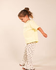 A young child wearing a pale yellow short-sleeve t-shirt and white flared pants with small dark star prints, standing against a plain beige studio background.