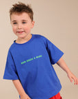 A young boy modeling a bright blue crewneck t-shirt with green text that reads 'HUN NEEDS A MARGI!' and red pants, standing against a solid beige studio background.