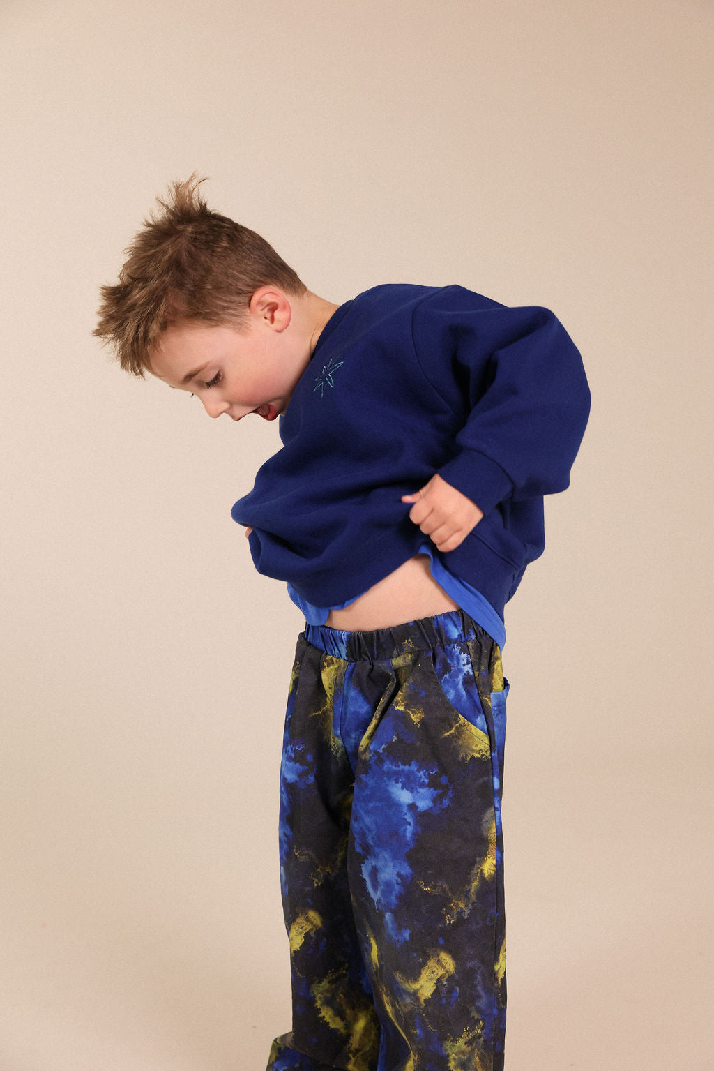 A young boy wearing a navy blue sweatshirt and blue and yellow patterned sweatpants, lifting his sweatshirt to show his midriff against a solid beige background.