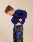 A young boy wearing a navy blue sweatshirt and blue and yellow patterned sweatpants, lifting his sweatshirt to show his midriff against a solid beige background.